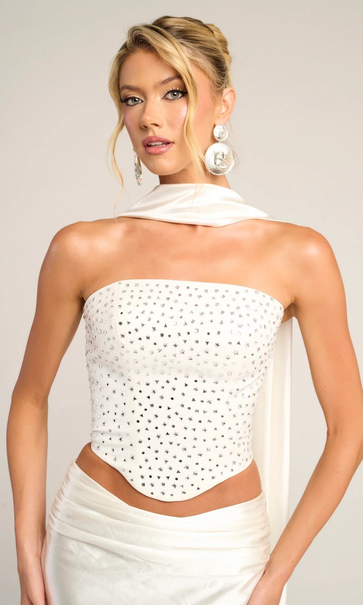 A woman wears the Portia and Scarlett PS26042 long formal dress featuring a white, beaded corset top with silver details, a matching satin skirt, large circular earrings, and a draped white scarf.